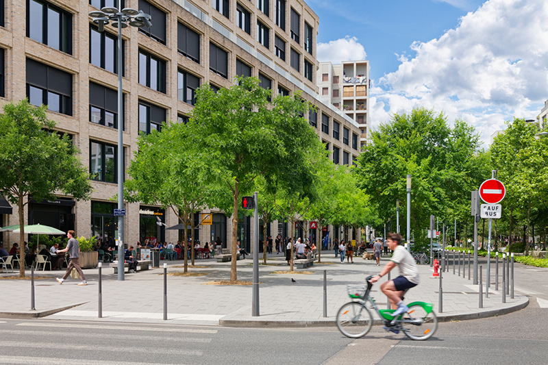 Photo d'une place où un vélo passe au premier plan et des piétons circulent devant les commerces et une terrasse